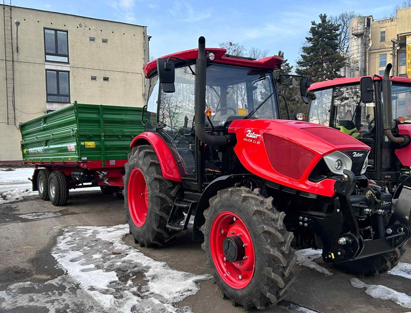 Na zdjęciu widać czerwony ciągnik rolniczy z podczepioną zieloną przyczepą, zaparkowany na placu z resztkami śniegu. Obok stoi drugi czerwony ciągnik. W tle są budynki i bezlistne drzewa, co wskazuje na zimę.